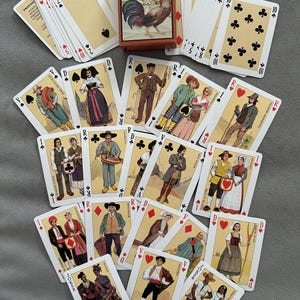 May include: A vintage deck of playing cards titled "Jeu des Provinces de France" featuring illustrations of people in traditional clothing. The card box shows a rooster. The cards are arranged on a gray surface.