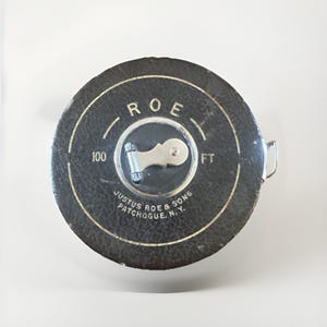 May include: A round, black, textured tape measure with a silver metal crank handle. The text reads ROE, 100 FT, and JUSTUS ROE & SONS PATCHOGUE, N.Y. The device features a circular design with a metallic center and a small metal pull tab on the side.