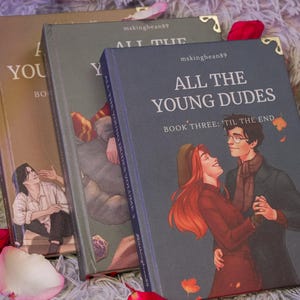 Puede incluir: Tres libros de tapa dura con ilustraciones y texto. El libro de la derecha se titula "ALL THE YOUNG DUDES" con "BOOK THREE: 'TIL THE END". Los otros libros tienen el texto "YOU" y "YOUNG DUDES VOLUME". Los libros son en tonos marr&oacute;n, gris y azul.