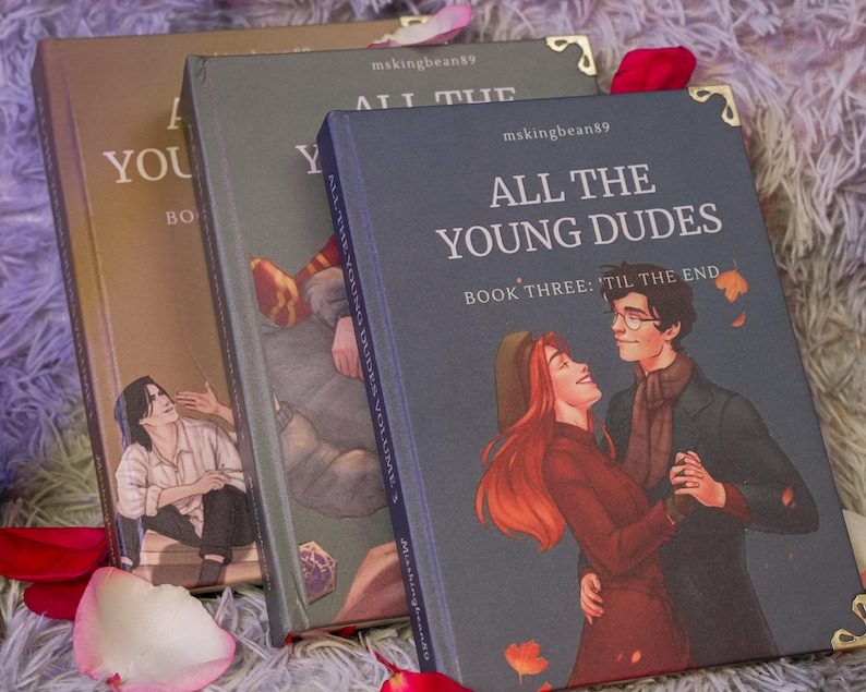 Puede incluir: Tres libros de tapa dura con ilustraciones y texto. El libro de la derecha se titula "ALL THE YOUNG DUDES" con "BOOK THREE: 'TIL THE END". Los otros libros tienen el texto "YOU" y "YOUNG DUDES VOLUME". Los libros son en tonos marr&oacute;n, gris y azul.