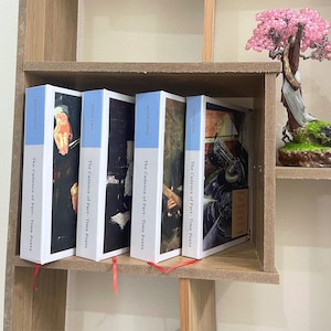 May include: Four books with white spines and light blue tops are displayed on a wooden shelf. The books are titled "The Cadence of Part-Time Poets" with "Book One", "Book Two", "Book Three", and "Book Four" visible. Red ribbons mark the pages.