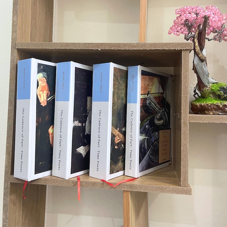 May include: Four books with white spines and light blue tops are displayed on a wooden shelf. The books are titled "The Cadence of Part-Time Poets" with "Book One", "Book Two", "Book Three", and "Book Four" visible. Red ribbons mark the pages.