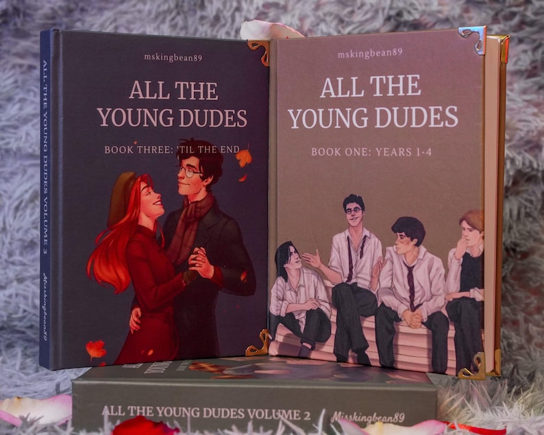 Puede incluir: Colecci&oacute;n de tres libros titulados "All The Young Dudes". Las portadas presentan ilustraciones de personajes y el texto "All The Young Dudes", "Book Three: 'Til The End" y "Book One: Years 1-4". Los libros est&aacute;n en un entorno decorativo.