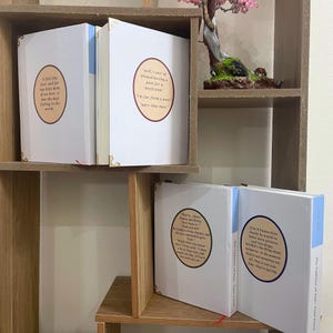 May include: White hardcover books with gold corner accents are displayed on a wooden shelf. Each book has a blue spine and a circular quote on the cover. The books are part of a collection.