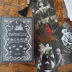May include: A black book titled "MANACLED" with white floral and snake illustrations, a crescent moon, and a castle. Several bookmarks and art prints with gothic illustrations and a black tassel are also visible.