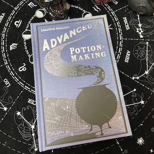 Op de afbeelding: Een hardcover boek getiteld "Advanced Potion-Making" door Libatius Borage. De kaft is lichtblauw met zilveren letters en een zilveren illustratie van een ketel. Het boek ligt op een zwarte ondergrond met een wit astrologisch ontwerp.