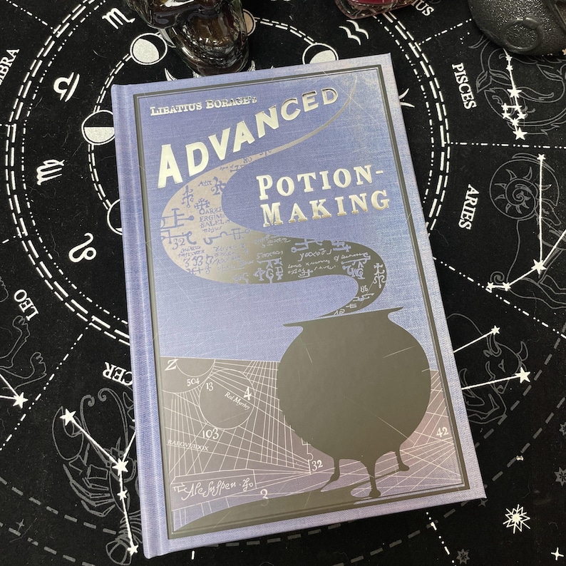 Op de afbeelding: Een hardcover boek getiteld "Advanced Potion-Making" door Libatius Borage. De kaft is lichtblauw met zilveren letters en een zilveren illustratie van een ketel. Het boek ligt op een zwarte ondergrond met een wit astrologisch ontwerp.