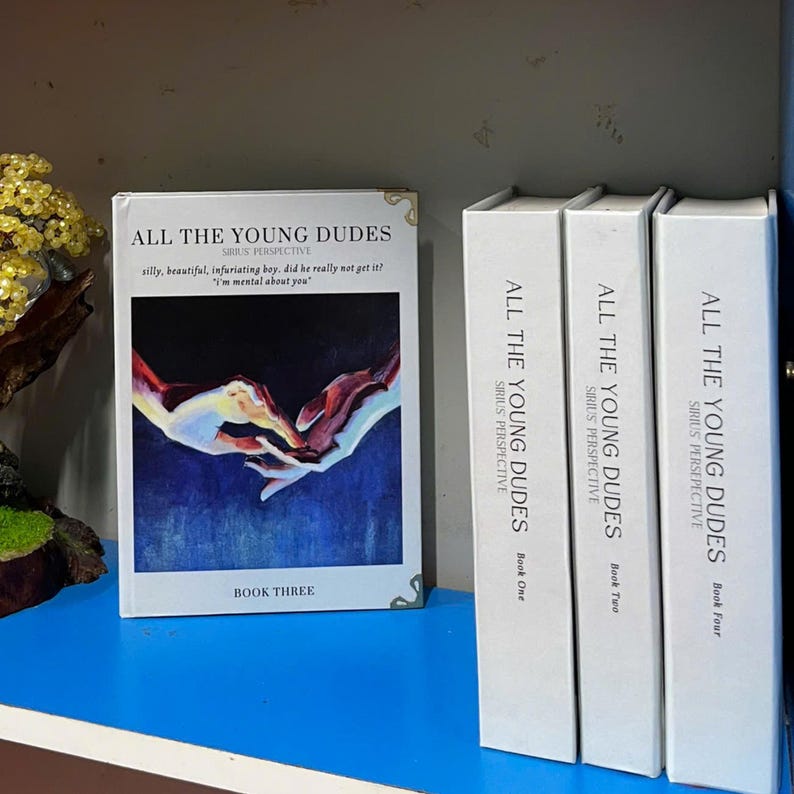 May include: A collection of books titled "ALL THE YOUNG DUDES" with white spines and matching covers. The book on the left is Book Three, featuring a colorful illustration of hands. The other books are Book One, Book Two, and Book Four.