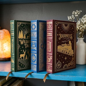 Puede incluir: Tres libros de tapa dura con títulos y diseños dorados en relieve. Los libros son en tonos verdes, azules y burdeos. Los títulos incluyen "Wilderness Tales", "Chronicles of Lost Realms" y "Journeys & Odysseys". Los libros están en una estantería azul.