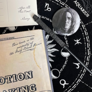 Op de afbeelding: Een zwarte toverstaf, een knoop met een portret en een boek getiteld "Advanced Potion Making" zijn gerangschikt op een zwarte astrologische kaart. Een ansichtkaart met de tekst "-After All This Time? -Always" is ook aanwezig. De items zijn thema's uit een fantasiewereld.