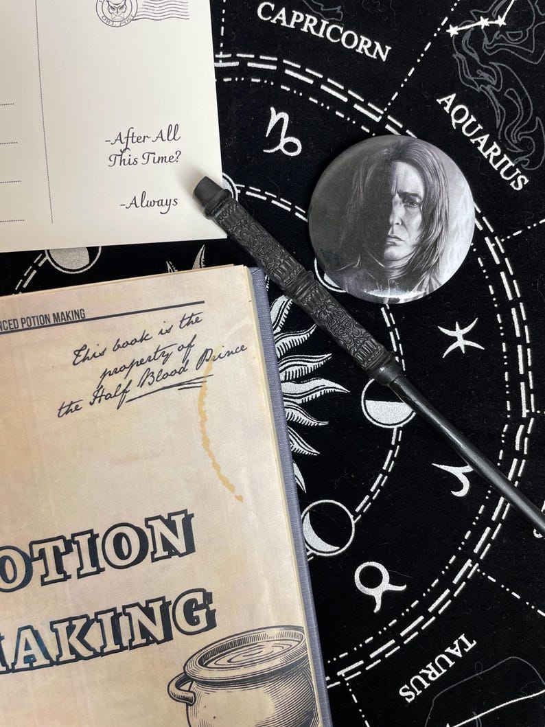Op de afbeelding: Een zwarte toverstaf, een knoop met een portret en een boek getiteld "Advanced Potion Making" zijn gerangschikt op een zwarte astrologische kaart. Een ansichtkaart met de tekst "-After All This Time? -Always" is ook aanwezig. De items zijn thema's uit een fantasiewereld.