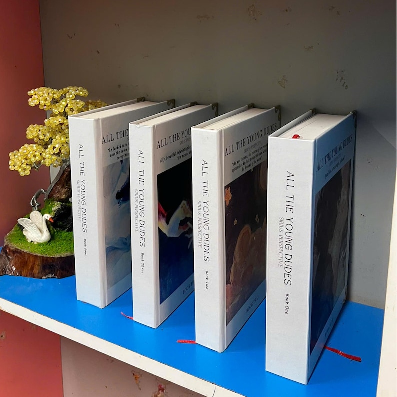 May include: Four white hardcover books titled "ALL THE YOUNG DUDES" with "SIRIUS PERSPECTIVE" on the spine. The books are arranged on a blue shelf. Book covers feature abstract art. A decorative display with a swan and yellow beads is in the background.