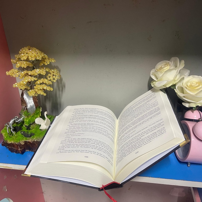 Puede incluir: Un libro abierto con p&aacute;ginas doradas y una cinta roja como marcador. Un &aacute;rbol decorativo con cuentas amarillas y una peque&ntilde;a escultura est&aacute;n a la izquierda. Rosas blancas y un despertador rosa est&aacute;n a la derecha.