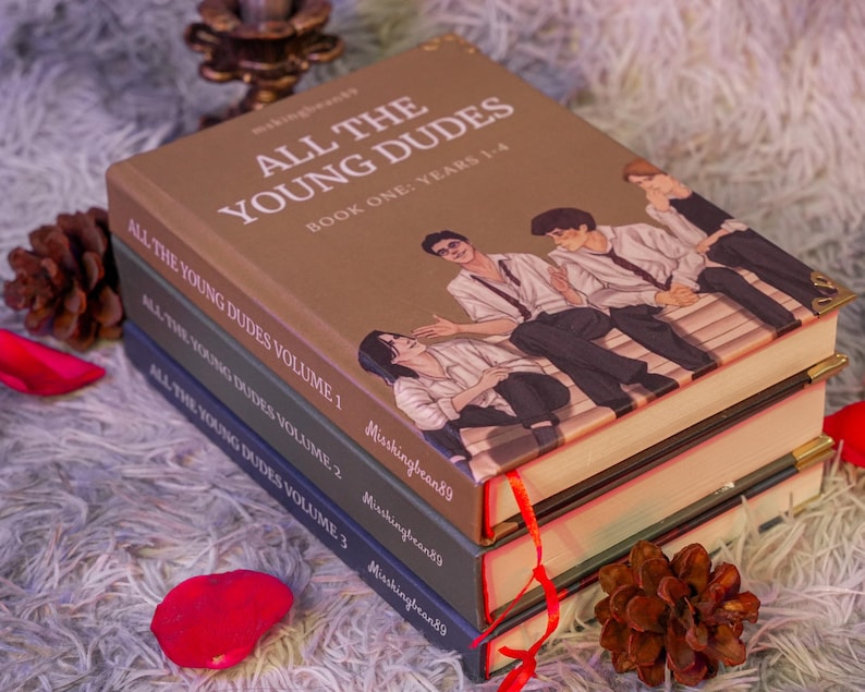 Puede incluir: Una pila de cuatro libros con el t&iacute;tulo "ALL THE YOUNG DUDES". El libro superior es marr&oacute;n con una ilustraci&oacute;n de j&oacute;venes. Los otros libros son de color verde oscuro y azul. Cintas rojas marcan las p&aacute;ginas.