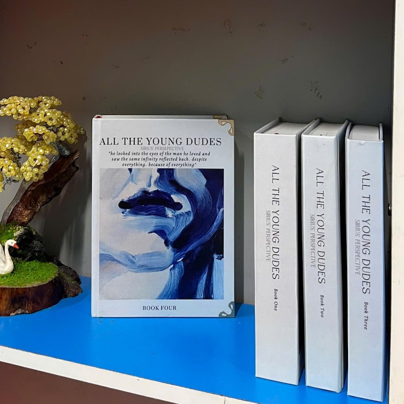 May include: A collection of books titled "ALL THE YOUNG DUDES" with a blue and white abstract design on the cover of Book Four. The spines of Books One, Two, and Three are visible. The books are on a blue shelf.