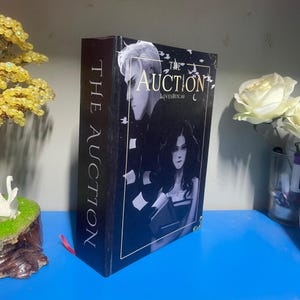 Puede incluir: Un libro de tapa dura titulado "The Auction" con una cubierta oscura que presenta ilustraciones de dos personas. El lomo dice "The Auction" verticalmente. El libro tiene un borde dorado y una cinta roja como marcador.