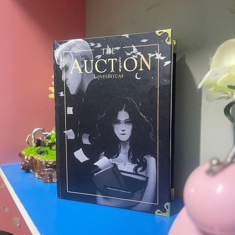 Puede incluir: Un libro de tapa dura titulado "The Auction" con una cubierta oscura que presenta ilustraciones de dos personas. El libro tiene protectores de esquina dorados y el texto "LOVESBITCAS" es visible. El fondo es un degradado de azul oscuro y negro.