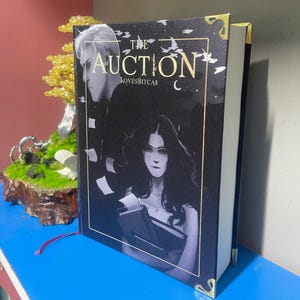 Puede incluir: Un libro de tapa dura titulado "The Auction" con una cubierta oscura que presenta ilustraciones de dos personas. El libro tiene protectores de esquina dorados y una cinta roja como marcador. El nombre del autor, LOVESBITCAS, tambi&eacute;n es visible.