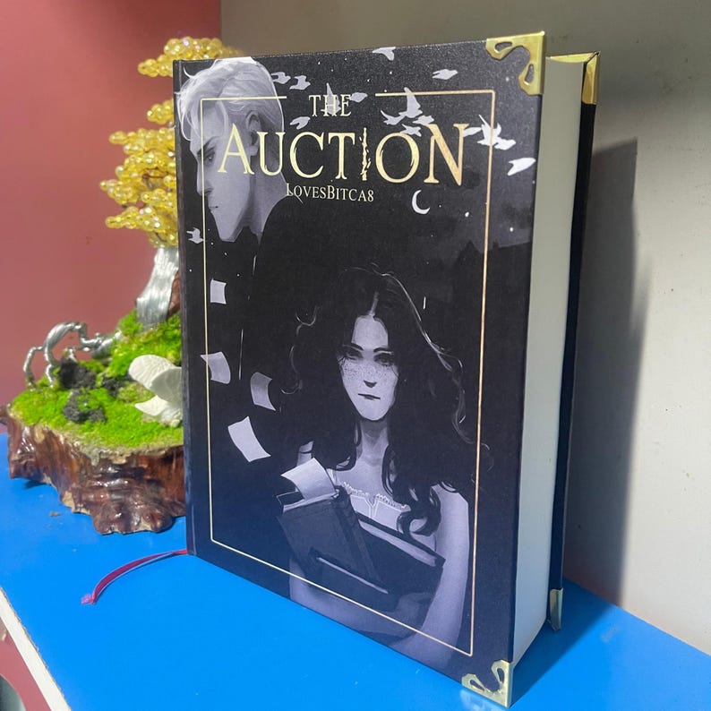 Puede incluir: Un libro de tapa dura titulado "The Auction" con una cubierta oscura que presenta ilustraciones de dos personas. El libro tiene protectores de esquina dorados y una cinta roja como marcador. El nombre del autor, LOVESBITCAS, tambi&eacute;n es visible.