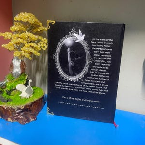 Puede incluir: Un libro de tapa dura negro con esquinas doradas y una cinta de lectura roja. La contraportada presenta texto y una imagen enmarcada. Un &aacute;rbol decorativo y una figura de p&aacute;jaro blanco est&aacute;n en el fondo.