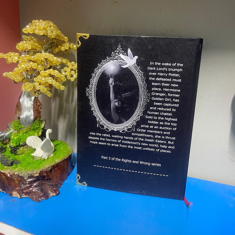 Puede incluir: Un libro de tapa dura negro con esquinas doradas y una cinta de lectura roja. La contraportada presenta texto y una imagen enmarcada. Un &aacute;rbol decorativo y una figura de p&aacute;jaro blanco est&aacute;n en el fondo.