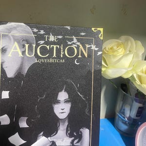 Puede incluir: Portada de libro con el t&iacute;tulo "The Auction" en letras doradas. La portada presenta un fondo oscuro con ilustraciones de dos personas. El texto "LOVESBITCAS" est&aacute; debajo del t&iacute;tulo. Un ramo de rosas amarillas est&aacute; en el fondo.
