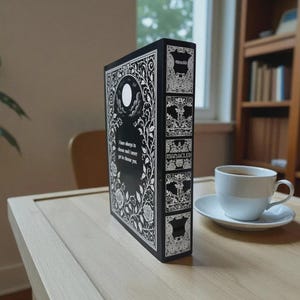 May include: A black book with white floral and moon designs, and the text "I have always to choose and I never get to choose you." The spine reads "MANACLEE" and "Ghoulish". A white teacup and saucer sit nearby.