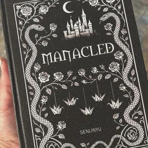 May include: A black book cover with the title "MANACLED" in white, featuring a castle, crescent moon, and snake and rose illustrations. The cover also includes origami cranes and the author's name, SENLINYU.