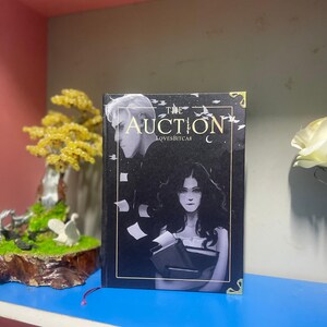 Puede incluir: Un libro de tapa dura titulado "The Auction" con una cubierta oscura que presenta ilustraciones de dos personas. El t&iacute;tulo est&aacute; en una fuente estilizada, y el nombre del autor, LOVESBITCAS, tambi&eacute;n es visible. El libro tiene esquinas doradas y una marca de libro roja.