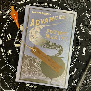 Op de afbeelding: Een boek getiteld "Advanced Potion-Making" met een lichtblauwe kaft en gouden letters. Een bruine leren bladwijzer is ingevoegd, met de tekst "After all this time? - Always." Een oranje glazen flesje en een toverstaf zijn ook zichtbaar.