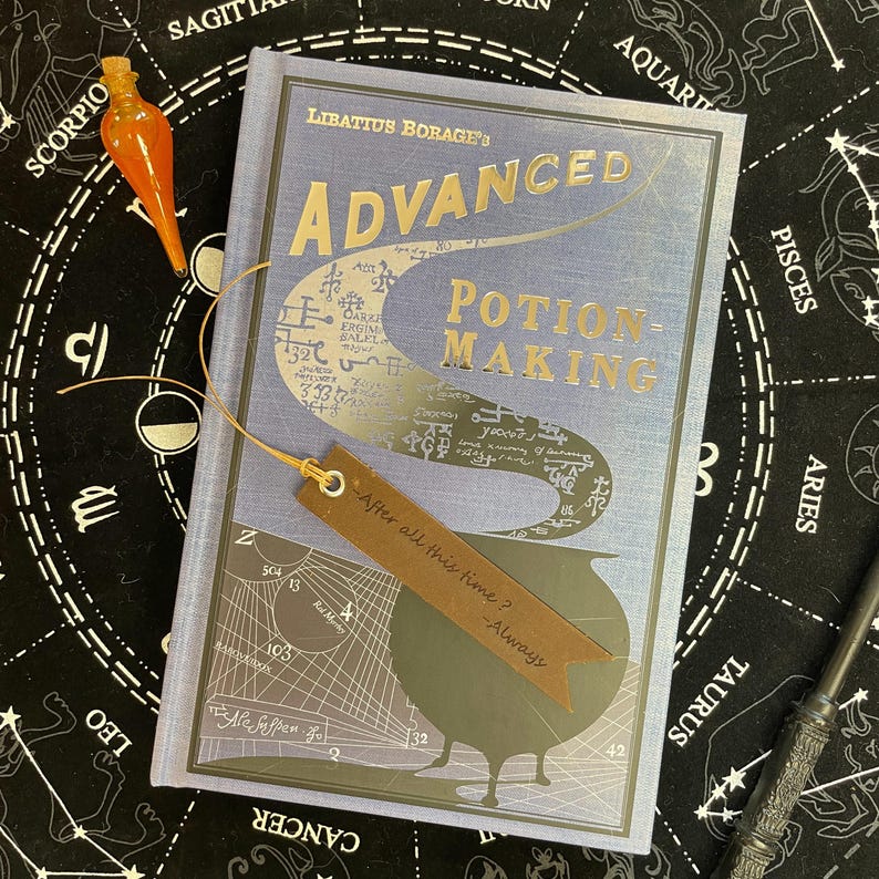 Op de afbeelding: Een boek getiteld "Advanced Potion-Making" met een lichtblauwe kaft en gouden letters. Een bruine leren bladwijzer is ingevoegd, met de tekst "After all this time? - Always." Een oranje glazen flesje en een toverstaf zijn ook zichtbaar.