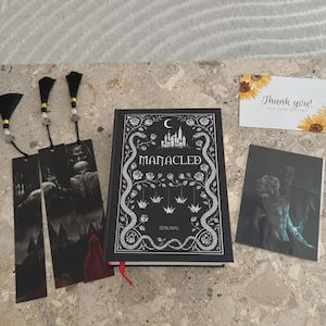 May include: A black book titled "MANACLED" with a white floral and snake design, accompanied by three black bookmarks with tassel ends and a small art print. A "Thank you!" card with sunflowers is also present.