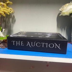 Puede incluir: Un libro negro titulado "THE AUCTION" con letras blancas. La portada presenta una imagen en escala de grises. El libro est&aacute; sobre una superficie azul, con elementos decorativos en el fondo.