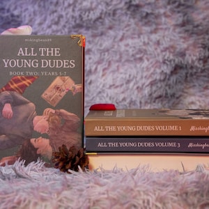 Puede incluir: Una pila de libros con el t&iacute;tulo "All The Young Dudes". El libro m&aacute;s grande se titula "Book Two: Years 5-7" y presenta una ilustraci&oacute;n de dos personas. Otros libros est&aacute;n etiquetados como "Volumen 1" y "Volumen 3". Los libros est&aacute;n sobre una superficie borrosa de color p&uacute;rpura claro.