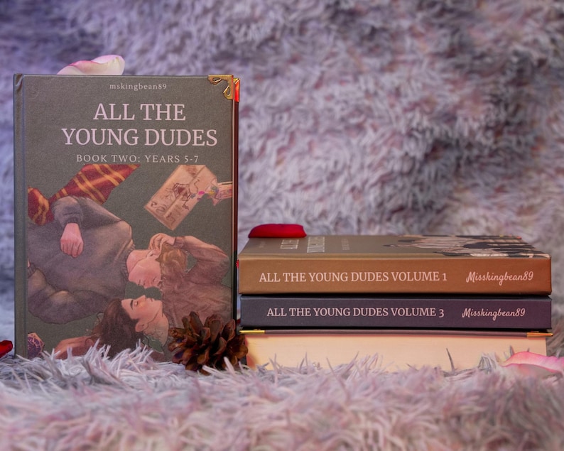 Puede incluir: Una pila de libros con el t&iacute;tulo "All The Young Dudes". El libro m&aacute;s grande se titula "Book Two: Years 5-7" y presenta una ilustraci&oacute;n de dos personas. Otros libros est&aacute;n etiquetados como "Volumen 1" y "Volumen 3". Los libros est&aacute;n sobre una superficie borrosa de color p&uacute;rpura claro.