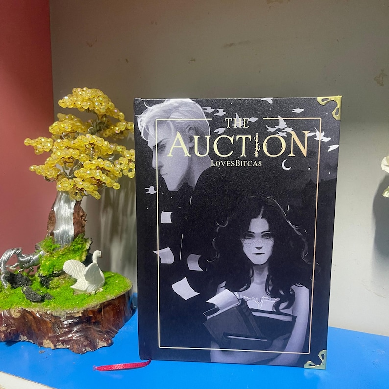 Puede incluir: Un libro titulado "The Auction" con una portada oscura que presenta dos figuras y el texto "LOVESBITCAS". El libro se muestra junto a un peque&ntilde;o &aacute;rbol decorativo con cuentas amarillas y una escultura en miniatura de un cisne.
