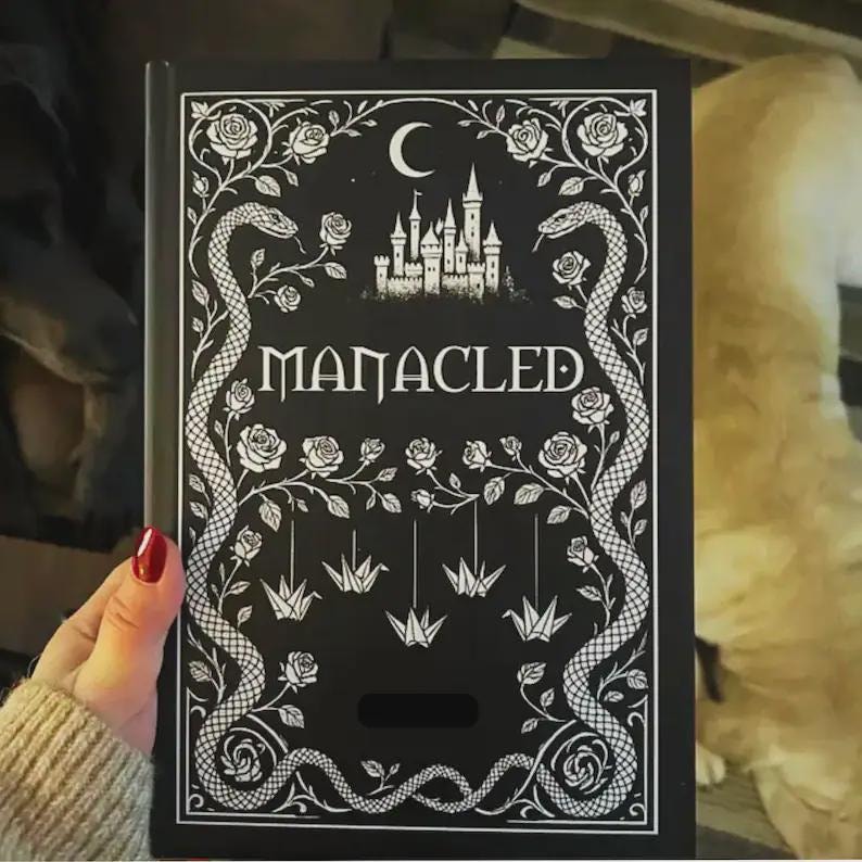 May include: A black book with the title "MANACLED" in white lettering. The cover features a white illustration of a castle, snakes, roses, a crescent moon, and origami cranes. The book is held by a person with red nail polish.