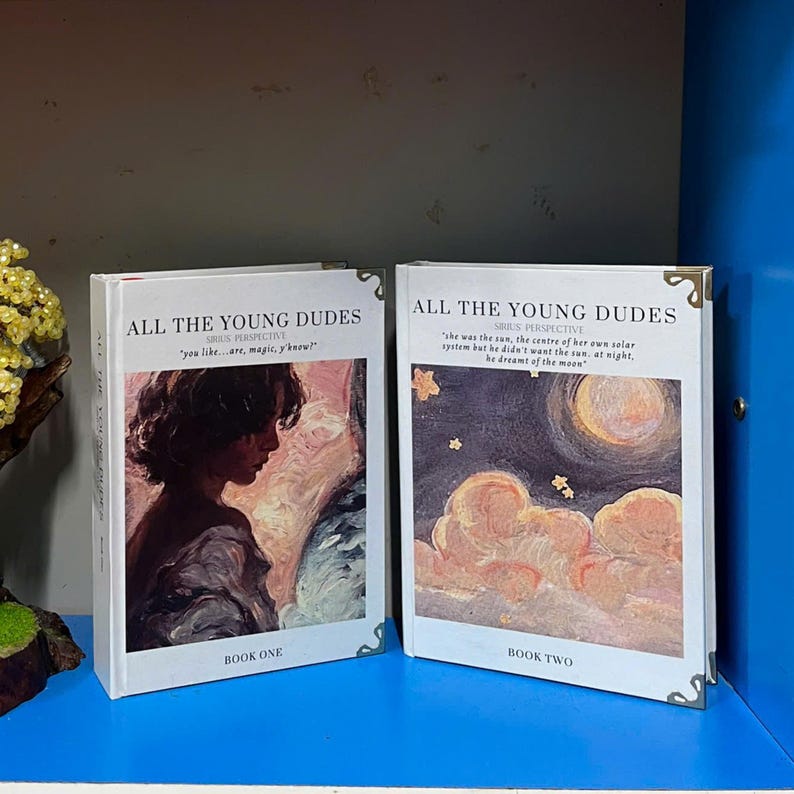 May include: Two hardcover books titled "ALL THE YOUNG DUDES" with decorative corner protectors. Book One features a profile portrait, while Book Two displays a night sky with a moon and clouds. The books have quotes and the text "BOOK ONE" and "BOOK TWO".