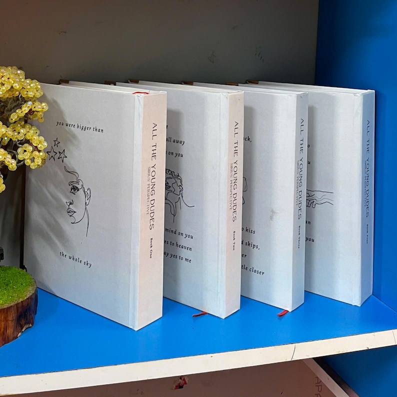 May include: A collection of white books with the title "ALL THE YOUNG DUDES" and various quotes and illustrations. The spines are white with the title and author's name. The books are on a blue shelf.