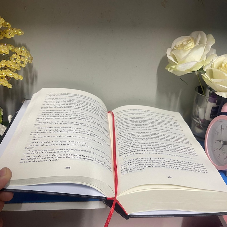 Puede incluir: Un libro abierto con p&aacute;ginas blancas y texto negro, con una cinta roja como marcador. El libro est&aacute; abierto, revelando el texto. Un jarr&oacute;n con rosas blancas y un despertador rosa est&aacute;n en el fondo.