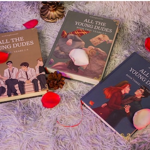 Puede incluir: Tres libros titulados "All The Young Dudes" se muestran sobre una superficie esponjosa, con p&eacute;talos de rosa rojos esparcidos. Las portadas de los libros presentan ilustraciones de personajes y el texto "Years 1-4", "Book Two: Years 1-7" y "Book Three: Til The End".