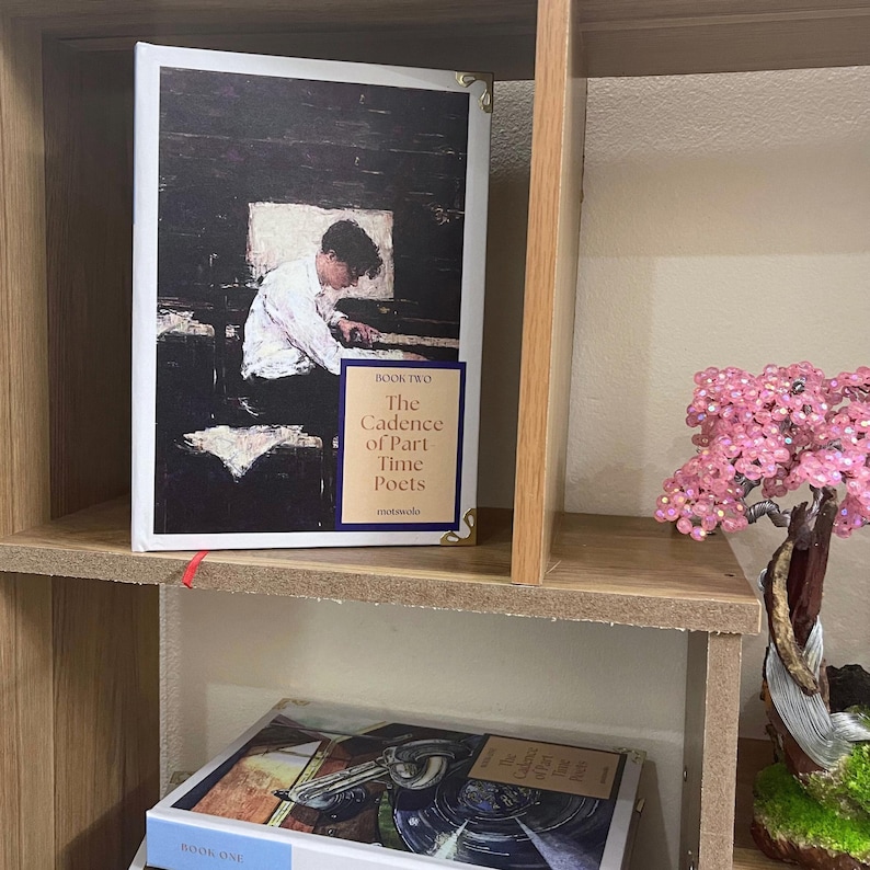 May include: A hardcover book titled "The Cadence of Part Time Poets" with a painting of a person at a piano. The book is on a wooden shelf with another book visible below. A small pink beaded tree is on the shelf.