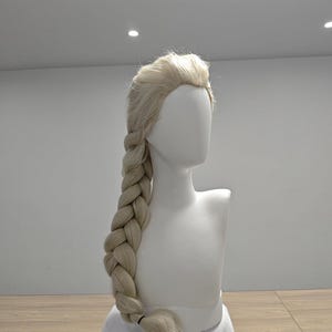 Op de afbeelding: Een platinablonde pruik gestyled met een volumineuze, dikke zijvlecht. Het haar is aan de voorzijde gescheiden met een lichte lift bij de kruin, en de lange, ingewikkelde vlecht hangt over de schouder, vastgezet met een klein zwart haarelastiekje.