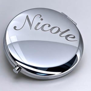 May include: A round, silver compact mirror with the name "Nicole" engraved on the top. The mirror has a shiny, reflective surface and a small clasp to open and close it. The compact is a classic and elegant accessory.