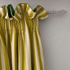 May include: A curtain featuring vertical stripes in olive green and cream. The fabric is gathered at the top with a ruffled header and hangs from a light gray metal rod with a pointed finial. The curtain has a solid dark green lining visible.