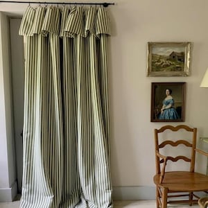May include: Floor-length curtains featuring a vertical striped pattern in olive green and off-white. The curtains are suspended from a black metal rod with rings and include a pleated valance at the top, creating a gathered, full-length drape.