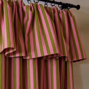 May include: A curtain featuring vertical stripes in alternating shades of olive green and bright pink. The fabric is gathered at the top and suspended from a black metal rod by a series of black circular rings, creating a pleated, draped effect.