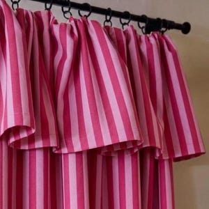 May include: A curtain featuring vertical stripes in alternating shades of hot pink and light pink. The fabric is gathered and suspended from a black metal rod by a series of black circular rings, creating a pleated effect across the top edge.