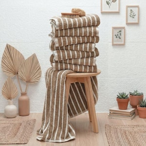 May include: A stack of seven folded brown and white striped towels resting on a light-colored wooden stool. A small wooden brush sits on top of the stack. The towels feature a consistent vertical stripe pattern throughout the fabric, creating a textured appearance.
