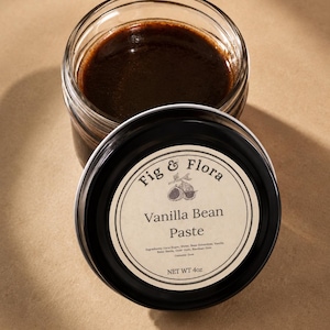 May include: A jar of Fig & Flora Vanilla Bean Paste with the lid slightly ajar. The jar contains a dark brown paste. The lid is black with a white label that reads "Fig & Flora Vanilla Bean Paste" and lists ingredients. Net weight is 4 oz.